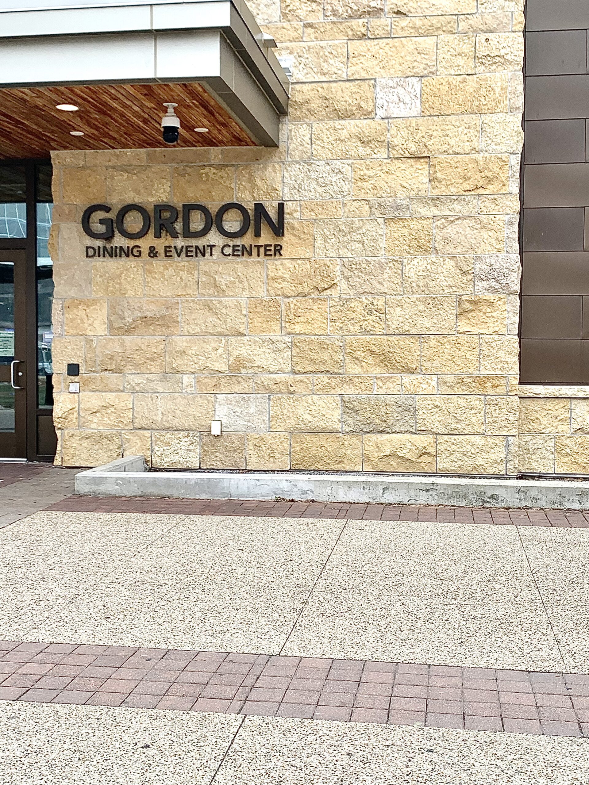 Gordon Dining & Event Center or Gordon Avenue Market. The picture is of Gordon's west entrance during a cloudy overcast day. Surrounding the perimeter of the marketplace is a concrete and brick walkway which leads to the black doors located on the west side of the building. The building is made of neutral-shaded bricks with wood and metal elements.