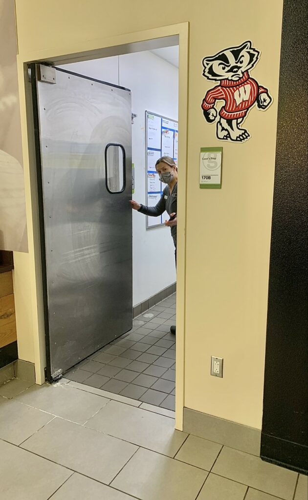 Sherman, the assistant director of dining, university dietician, and UW-Madison alumni. Sherman is a white woman with mid-length brown hair. She is wearing a grey sweater and black dress pants. Her arm is extended holding a large, stainless-steel door open. The door leads to the back of the market where the 'cook's prep' is located.