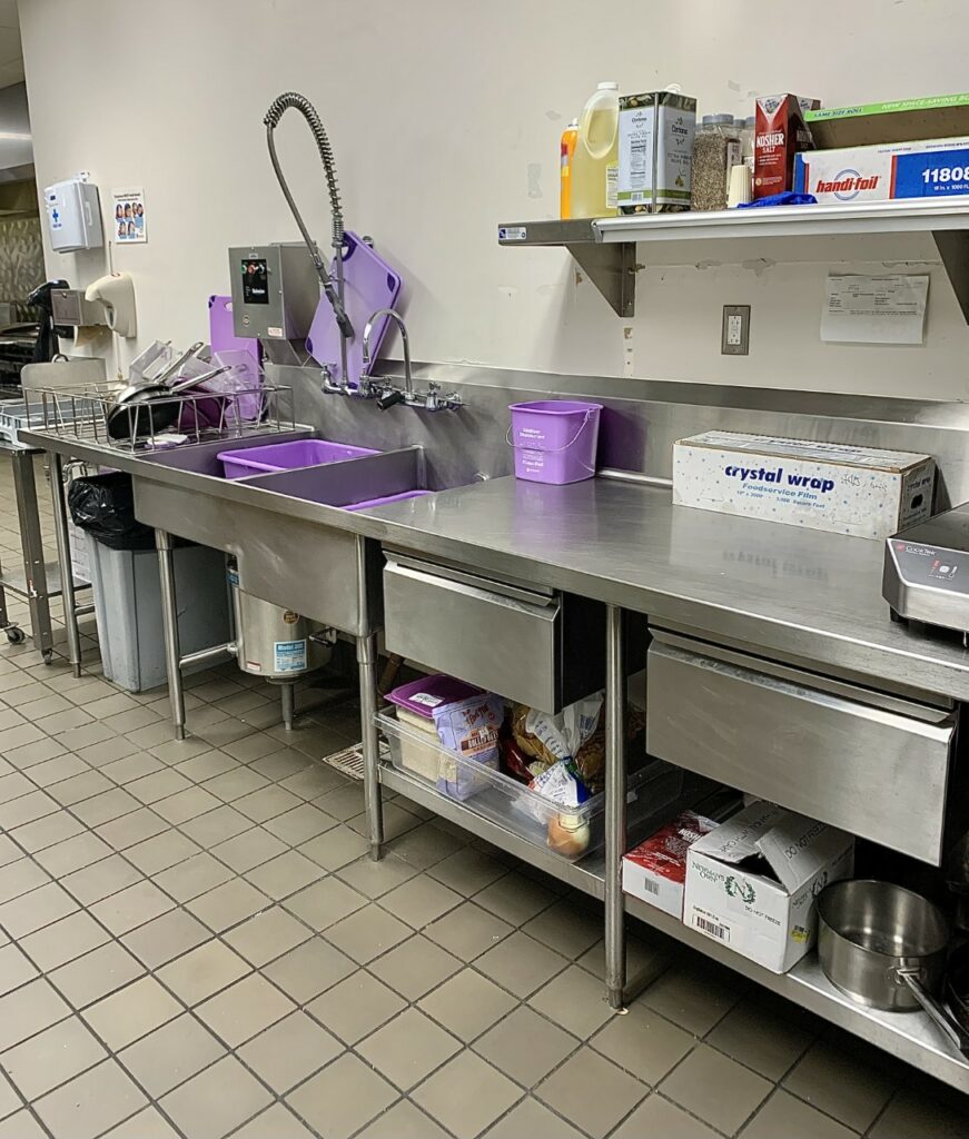 The "purple" station in the back of the ‘chef’s prep’ inside Gordon Avenue Market. The purple station kitchen space features a stainless steel counter, shelves, and sinks. Splayed around the kitchen are various allergen-free cooking ingredients and designated all-purple cooking utensils.