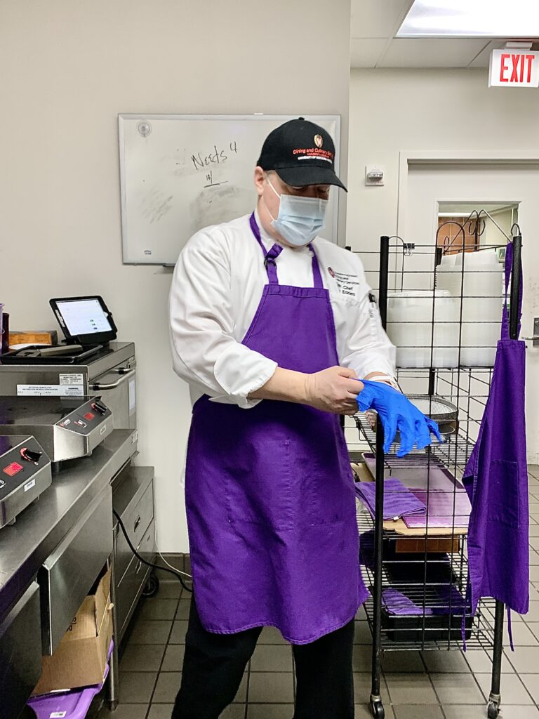 Kris Eckles, the head chef at Gordon Avenue Market preparing to cook an allergen-free meal in the “purple” station. Eckles is in chef's attire which includes a white, button-up shirt, black dress pants, and a black 'Dining and Culinary Services' cap. He has on a purple apron to designate he is in an allergen-free work space. He is demonstrating proper food-safety techniques by putting on blue, latex gloves.