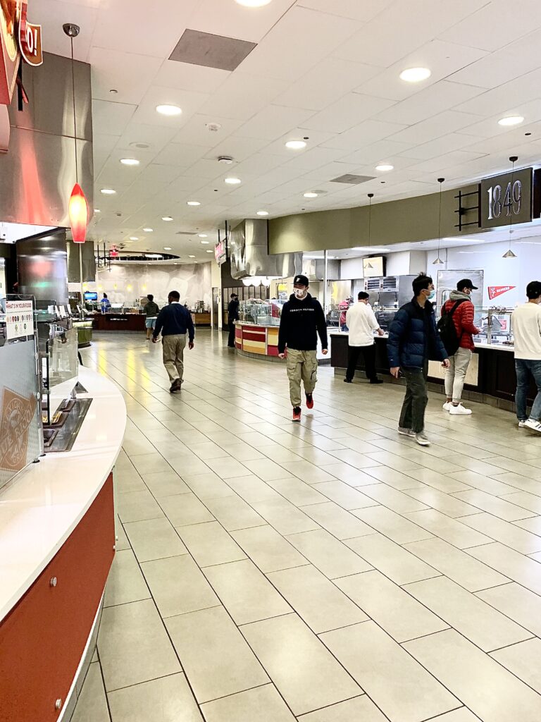 Students and staff walking the aisles of venues during lunchtime at Gordon Avenue Market. The main walkway of Gordon's has beige tile floors and is dimly lit with warm lights. On the right-side aisle are food venues 1849 and Eggcetera, and visible on the left side is Qué Rico.