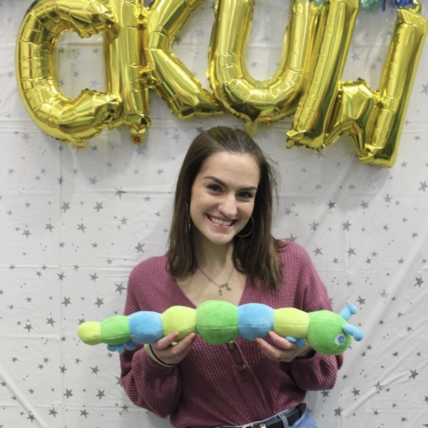 Kinzie Collins is standing in front of a blank white sheet with silver starts and big golden balloons that spell out CKUW (Camp Kesem University of Wisconsin). She is smiling holding a stuffed caterpillar (the logo for Camp Kesem) wearing a purple top and blue jeans.