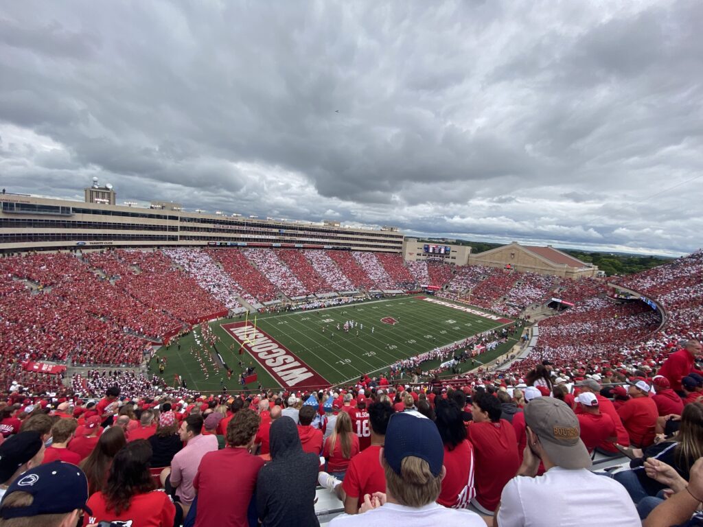 A view from above Camp Randall Stadium from the upper level stands. The stands are filled with red and white stripes. The sky is cloudy.