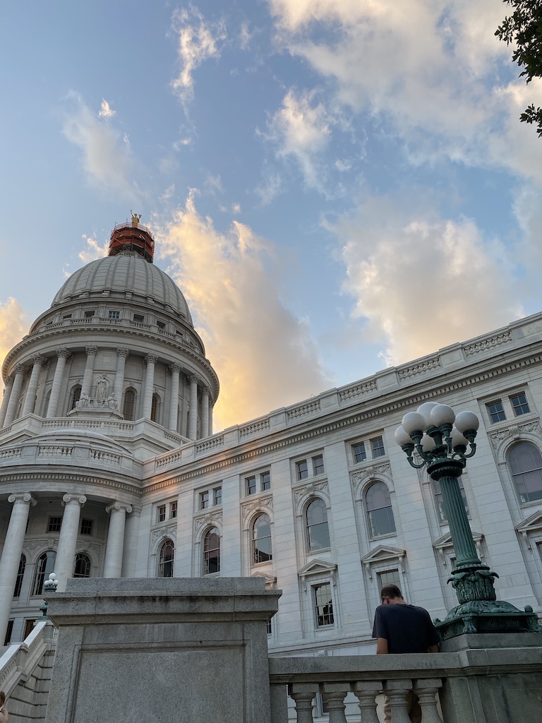 Lower angle photo of Capitol building with pink cloudss on the blue sky. There is a man with his back facing the camera in the lower right hand corner