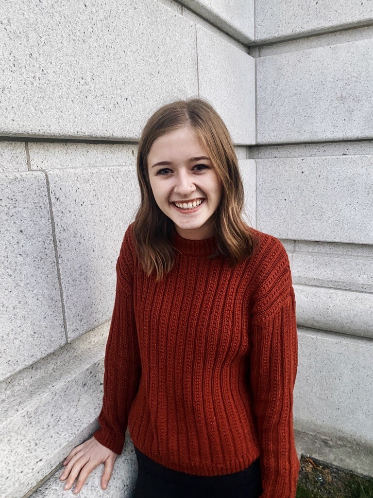 Brelynn Bille is outside and standing next to a light gray wall. She is a white woman with brown hair that is shoulder-length and in the image she is smiling. She is wearing a reddish-orange sweater and her hand is resting on a gray ledge connected to the wall she is next to.  